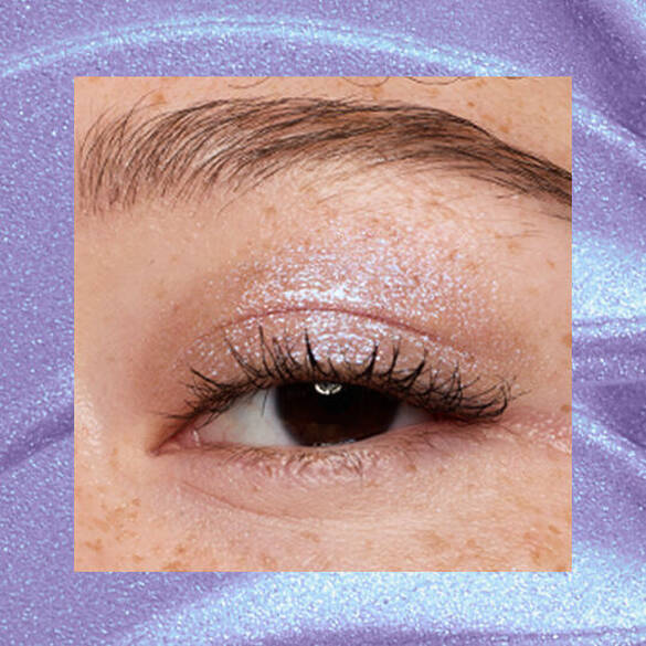 Close-up of an eye area with NYX Professional Makeup Lid Lingerie Cream Eye Pigment in 10 Lacy Lilac, shown on a duochrome blue textured background.