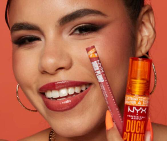 A smiling woman holds a NYX lip plumper and lip pencil against a vibrant orange background.