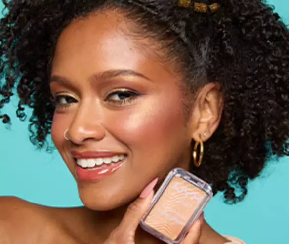 A smiling woman with curly hair holds a blush compact, showcasing her radiant makeup against a bright turquoise background.
